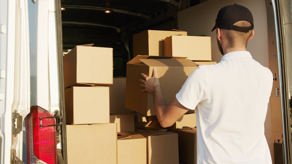 A man wearing a black cap and a white polo shirt is inside a moving van, loading brown cardboard boxes onto a shelf or stack. Several boxes of various sizes are arranged inside the van, some stacked o