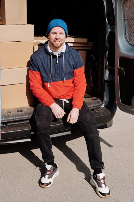 A man wearing a black cap and a white polo shirt is inside a moving van, loading brown cardboard boxes onto a shelf or stack. Several boxes of various sizes are arranged inside the van, some stacked o