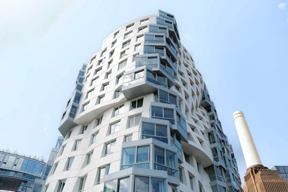 A red double-decker bus is traveling along a city street in London, passing modern office buildings with large glass windows and a classical-style stone facade with ornamental carvings. Pedestrians ar