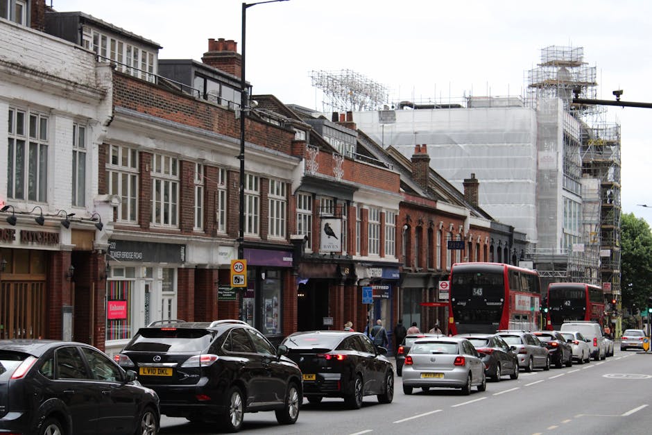 View of a street scene near Elephant and Castle with a row of traditional brick buildings and shops on the left, and a large building under scaffolding on the right. Several cars, including a black SUV, a silver hatchback, and other smaller vehicles, are parked along the curbside, with some in motion. Two red double-decker buses are visible, heading along the road. In the foreground, a parking lane is occupied by various cars, and pedestrians walk along the pavement near shop entrances. The scene is set during daylight hours under overcast skies, with urban surroundings indicating an active area for house removals or furniture transport, as professional moving services from companies like manandvanelephantandcastle.com support local home relocations and moving logistics.