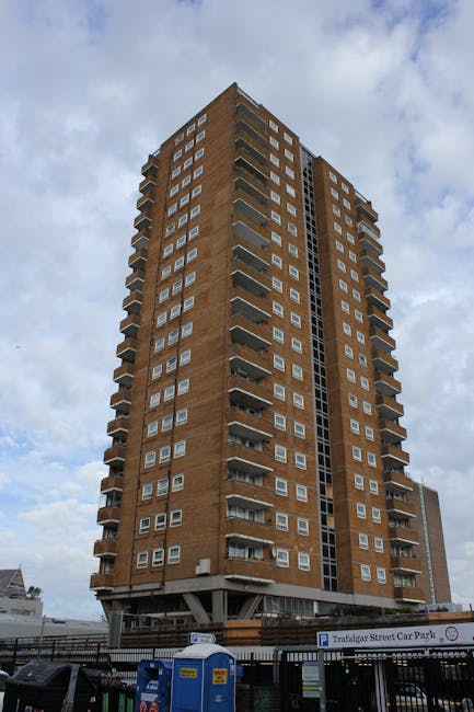 A tall, multi-storey residential building constructed from reddish-brown brick, with numerous balconies featuring metal railings on each floor, situated in an urban area. The building has a central vertical section with glass panels, and the sky above is partly cloudy. In the foreground, a vehicle parking area includes a sign indicating 'Trafalgar Street Car Park' and various parked cars, with some visible cardboard boxes, plastic wrapping, and furniture blankets at the base of the building, suggesting recent or ongoing home relocation activities. The environment appears well-lit with natural daylight, and the scene reflects typical urban house removals and furniture transport logistics managed by [COMPANY_NAME].