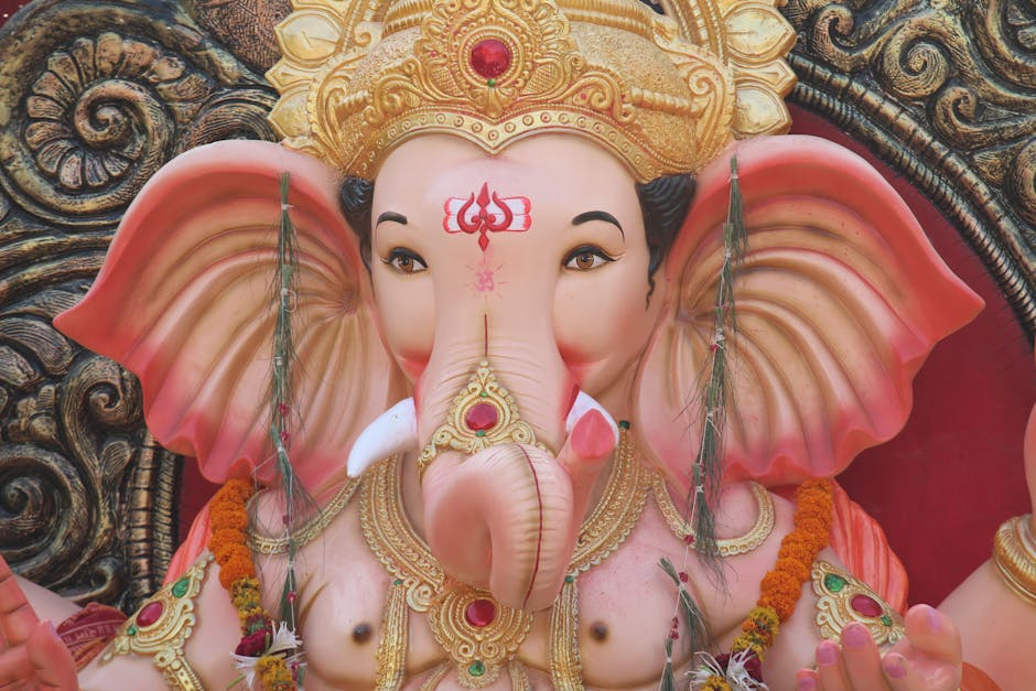 Close-up of a large, detailed statue of Lord Ganesha, the Hindu deity with an elephant head, adorned with intricate gold jewelry, a golden crown, and a red gemstone. The statue’s ears are pink with layered folds, and the face has a gentle expression with painted eyebrows, eyes, and a tilak on the forehead. The statue is decorated with flower garlands, and the background features metallic and ornamental patterns. This image depicts a religious or cultural display, with the statue likely positioned indoors or in a temple setting, not related to house removals or moving services. Occasionally, [COMPANY_NAME] offers packing and moving solutions, including handling delicate religious or cultural artefacts during home relocations.