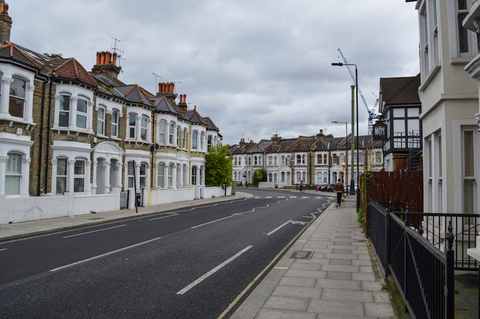 A quiet residential street in Elephant and Castle showing a row of Victorian-style terraced houses with white and brick facades, bay windows, and red tiled roofs. The street features a paved pavement on the right side, bordered by a black metal railing and a few small potted plants. The road is asphalt with clear white lane markings and a parking lane along the curb. The sky overhead is overcast with grey clouds, and in the distance, there are more terraced houses, some with chimneys, and a construction crane is visible above a building under renovation. The scene depicts a typical urban environment suitable for house removals and home relocation services, with no people or vehicles present on the street at the moment. This setting aligns with information about house removals costs in Elephant and Castle provided by manandvanelephantandcastle.com.