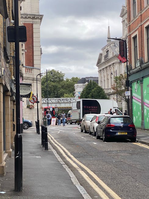 A street scene near the Elephant and Castle area showing vehicles parked along the curb, including a white moving van positioned for loadings, with several cardboard boxes and wrapped furniture on its flatbed. The pavement has black bollards separating pedestrian walkway from the roadway. In the background, people are gathered at a street entrance, with some carrying items, possibly related to home relocation or furniture transport. Buildings on both sides include a green storefront, a red brick structure, and historic architecture with a prominent white building topped with a spire visible under a cloudy sky. This scene depicts a typical urban environment suitable for house removals and moving services, with the presence of packing materials, loading activity, and parking arrangements aligning with Southwark Council parking rules for same-day removals.