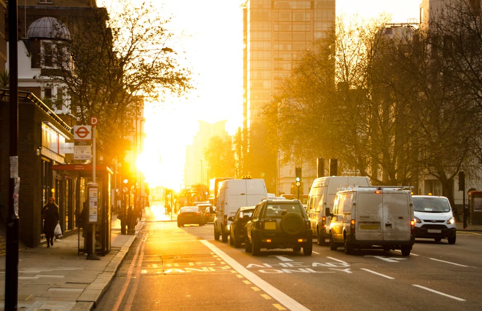 A busy urban street scene during sunset in Southwark, London, featuring a row of vehicles including white delivery vans, small cars, and a black car on a multi-lane road. The vehicles are parked and queued along the roadside, with some actively being loaded or unloaded as indicated by the presence of a few people on the busy pavement nearby. The sidewalk includes a bus stop shelter with a red London transport sign, and pedestrians walking past. Tall modern office buildings and older architectural structures line the background, partially silhouetted against the warm, golden sunlight filtering through the buildings and trees with bare branches. The scene captures the typical environment of a busy city without obstruction, illustrating the logistics involved in home relocation and furniture transport, relevant to Southwark Council parking rules for same-day removals, as seen on manandvanelephantandcastle.com.