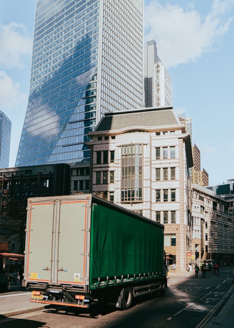 A street scene near the Elephant and Castle area showing vehicles parked along the curb, including a white moving van positioned for loadings, with several cardboard boxes and wrapped furniture on its flatbed. The pavement has black bollards separating pedestrian walkway from the roadway. In the background, people are gathered at a street entrance, with some carrying items, possibly related to home relocation or furniture transport. Buildings on both sides include a green storefront, a red brick structure, and historic architecture with a prominent white building topped with a spire visible under a cloudy sky. This scene depicts a typical urban environment suitable for house removals and moving services, with the presence of packing materials, loading activity, and parking arrangements aligning with Southwark Council parking rules for same-day removals.